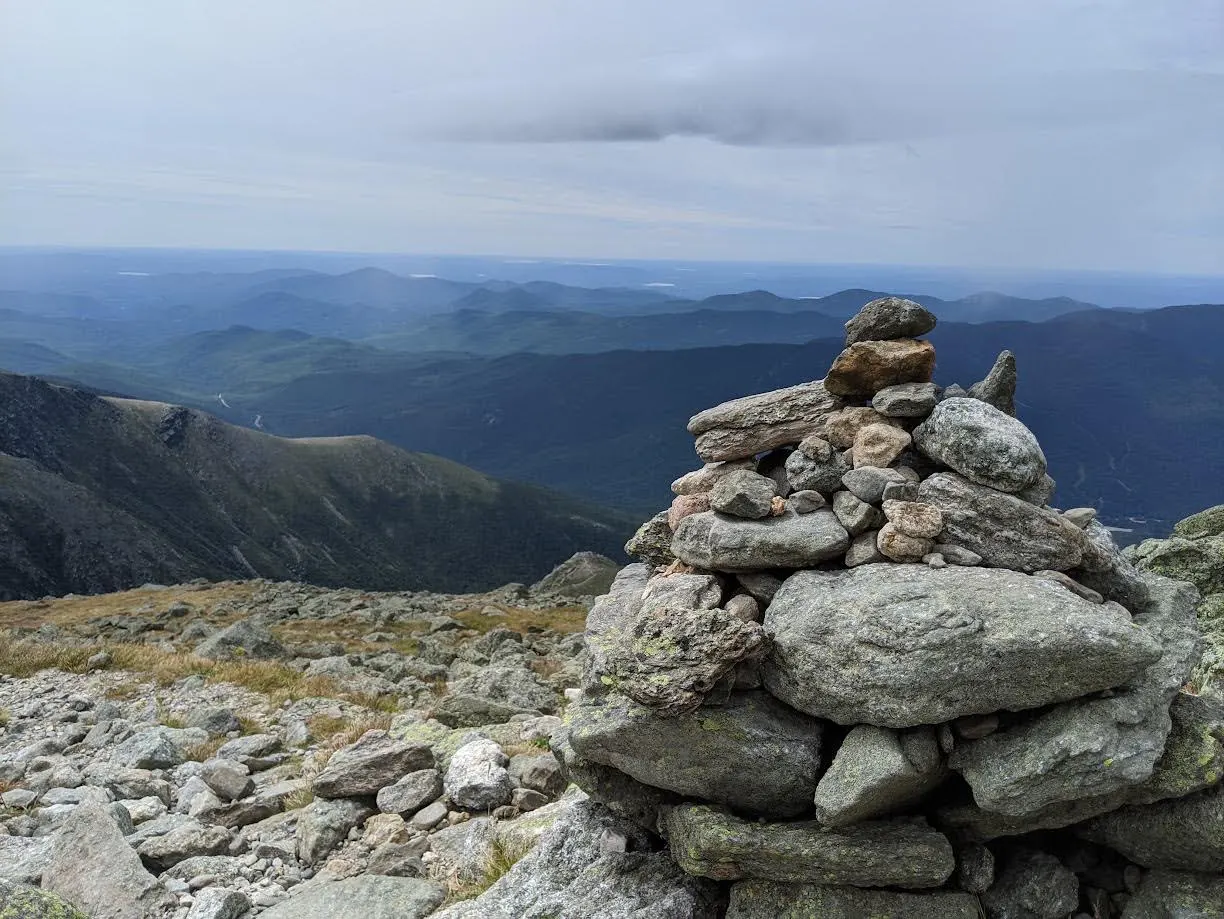 A stacked stone cairn on a rocky mountain summit above layered green ridges under an overcast sky